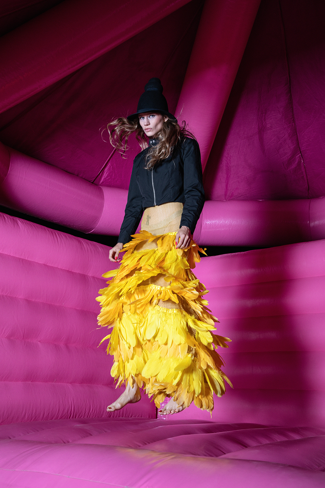 Model in bright yellow dress and black jacket posing mid-air in a bright pink bounce house